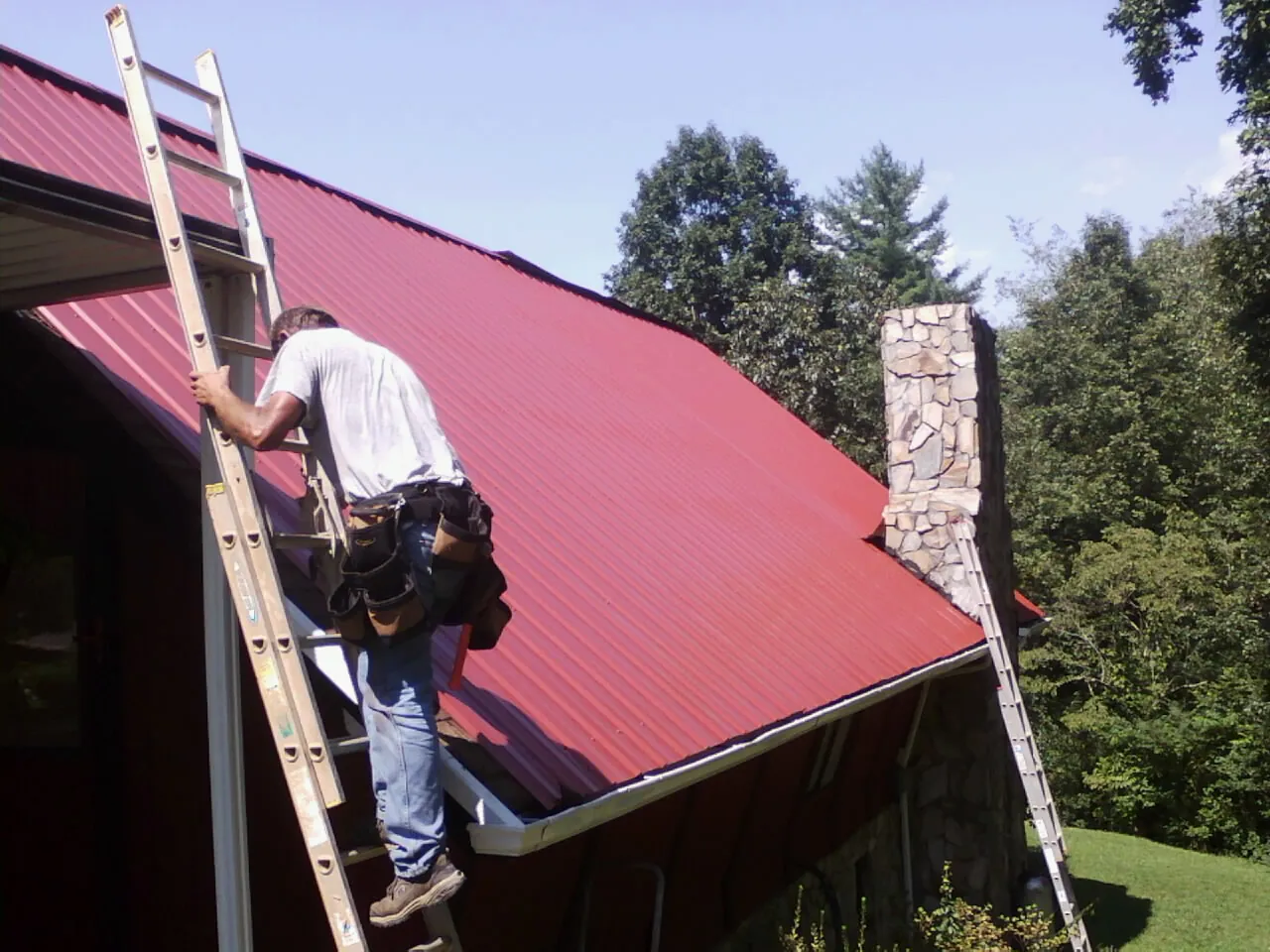 Roofer on a ladder beside a red metal roof and stone chimney — Soffit & Fascia Repair by Roofing Brothers in West Chesterfield